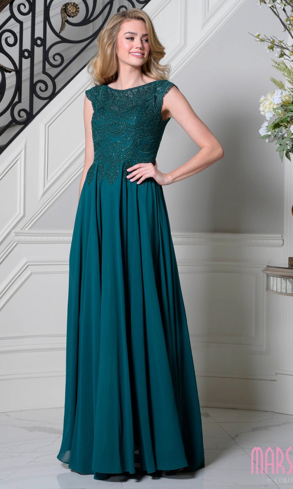 A woman in the Marsoni M238 Long Formal Dress in deep green, featuring lace detailing, stands indoors by a staircase, smiling and posing with one hand on her hip.